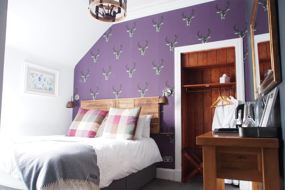 Un dormitorio con cama, cabecera de madera, pared morada con papel tapiz de ciervos y un armario con puertas de madera, además de una mesita de noche con lámpara.