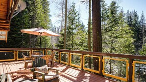 Terraza de madera con mobiliario exterior, una fogata y vistas a un bosque.