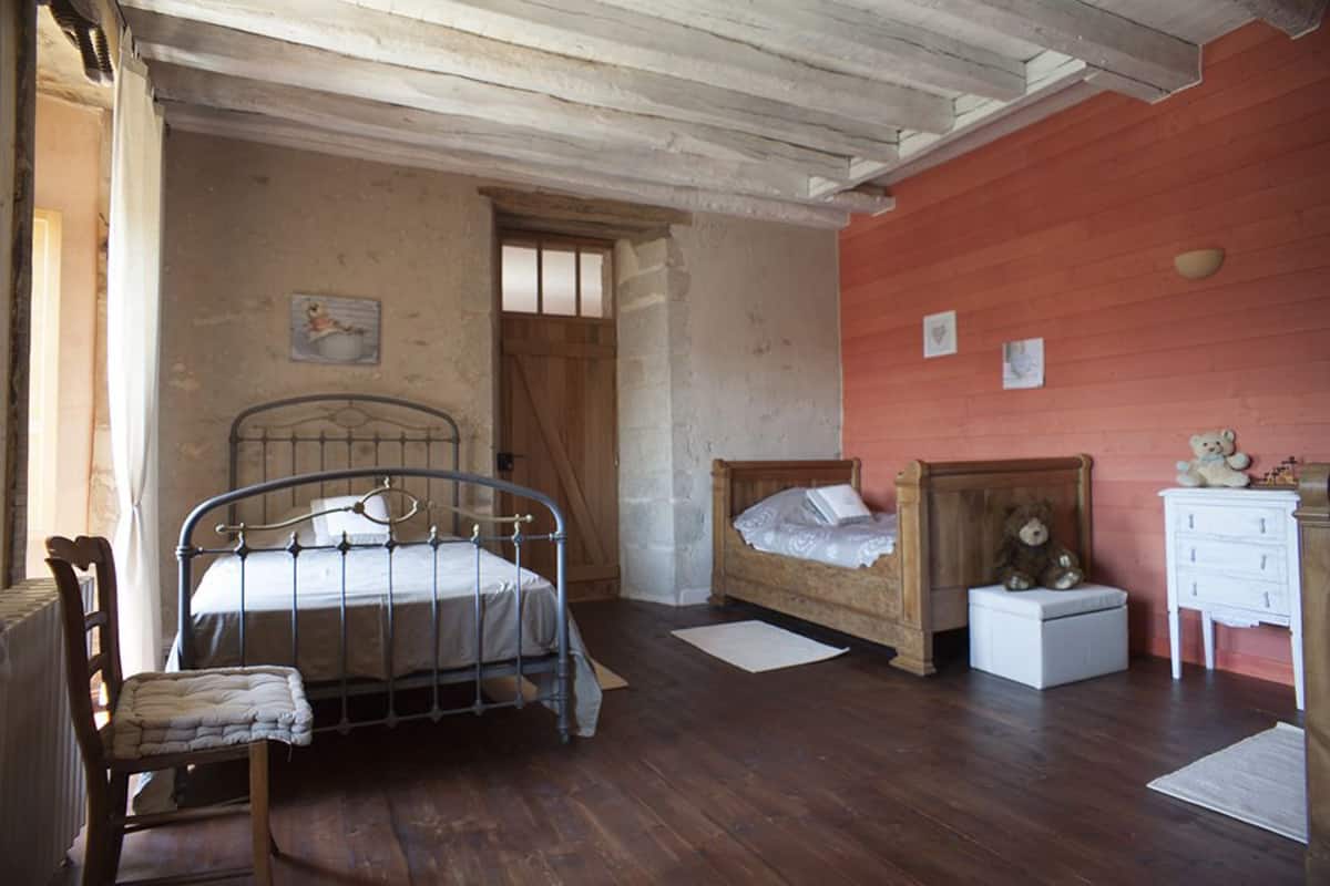Un dormitorio con estructura de cama metálica, cama de madera, una silla, una mesita de noche con un osito de peluche y un aparador blanco.