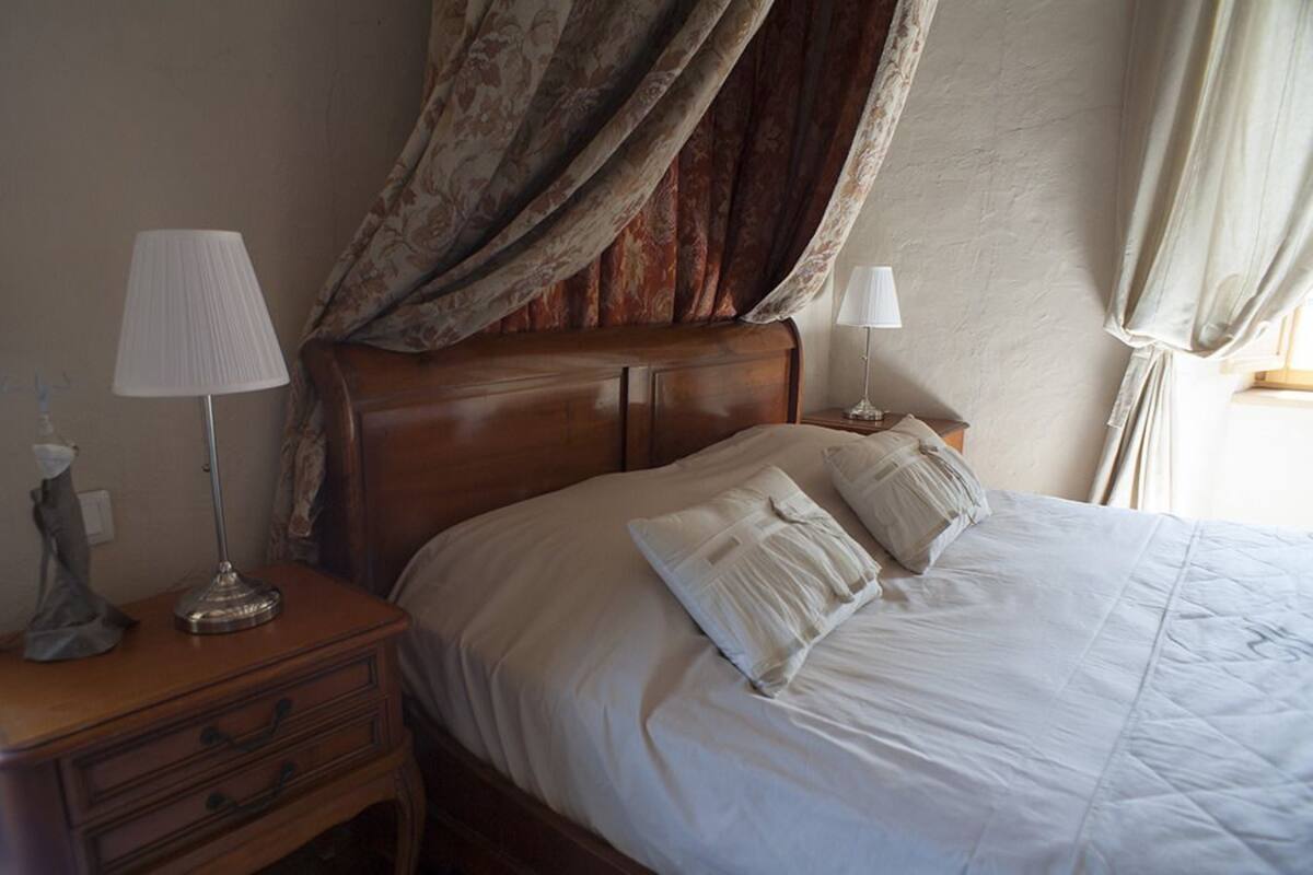 Una cama bien hecha con sábanas blancas y dos almohadas, una mesita de noche de madera con una lámpara y un objeto decorativo a la izquierda.