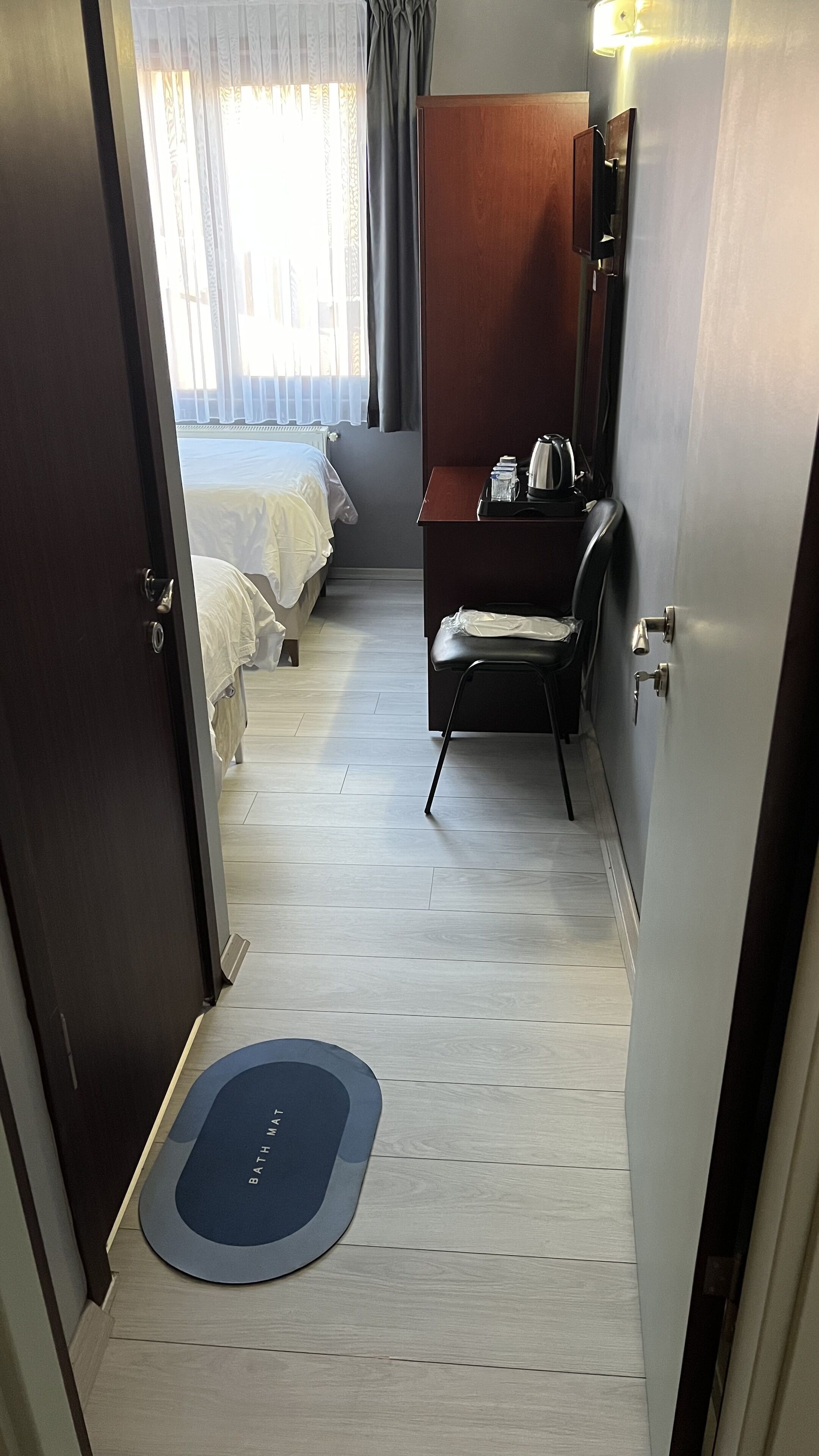 A hotel room with a bed, a desk with a chair, a desk lamp, and a bedside table with a kettle.