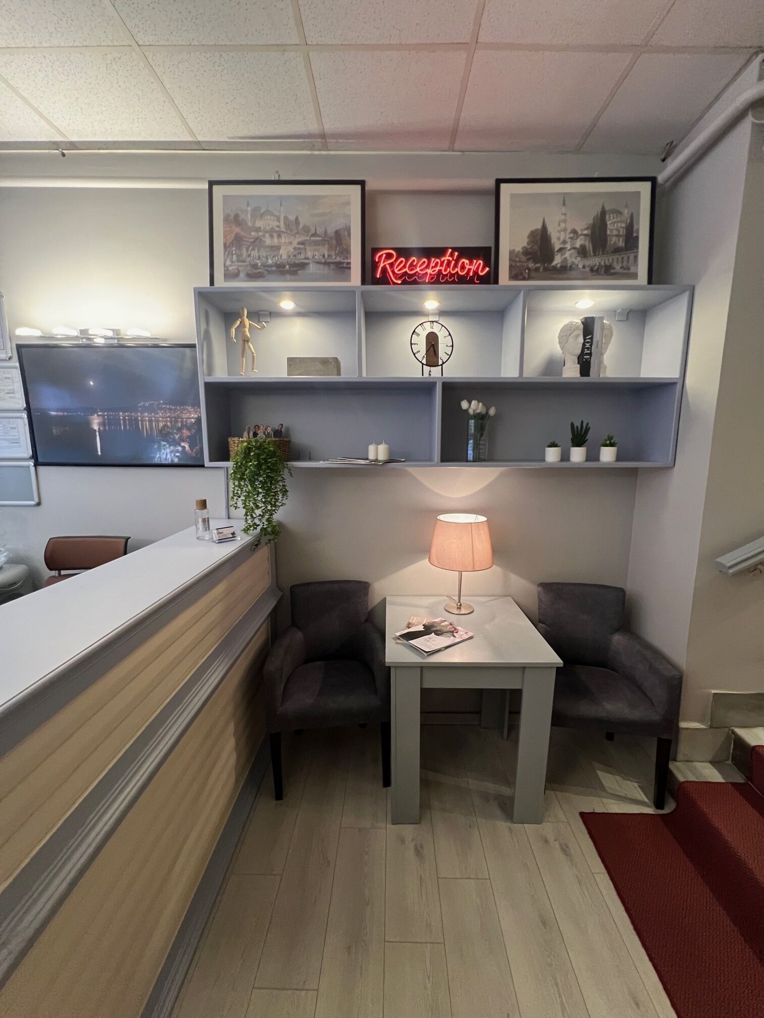 A reception area with a reception desk, two armchairs, a table with a lamp, a clock, and framed pictures on shelves.