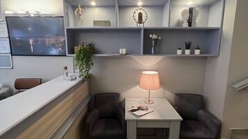 A reception area with a reception desk, two armchairs, a table with a lamp, a clock, and framed pictures on shelves.