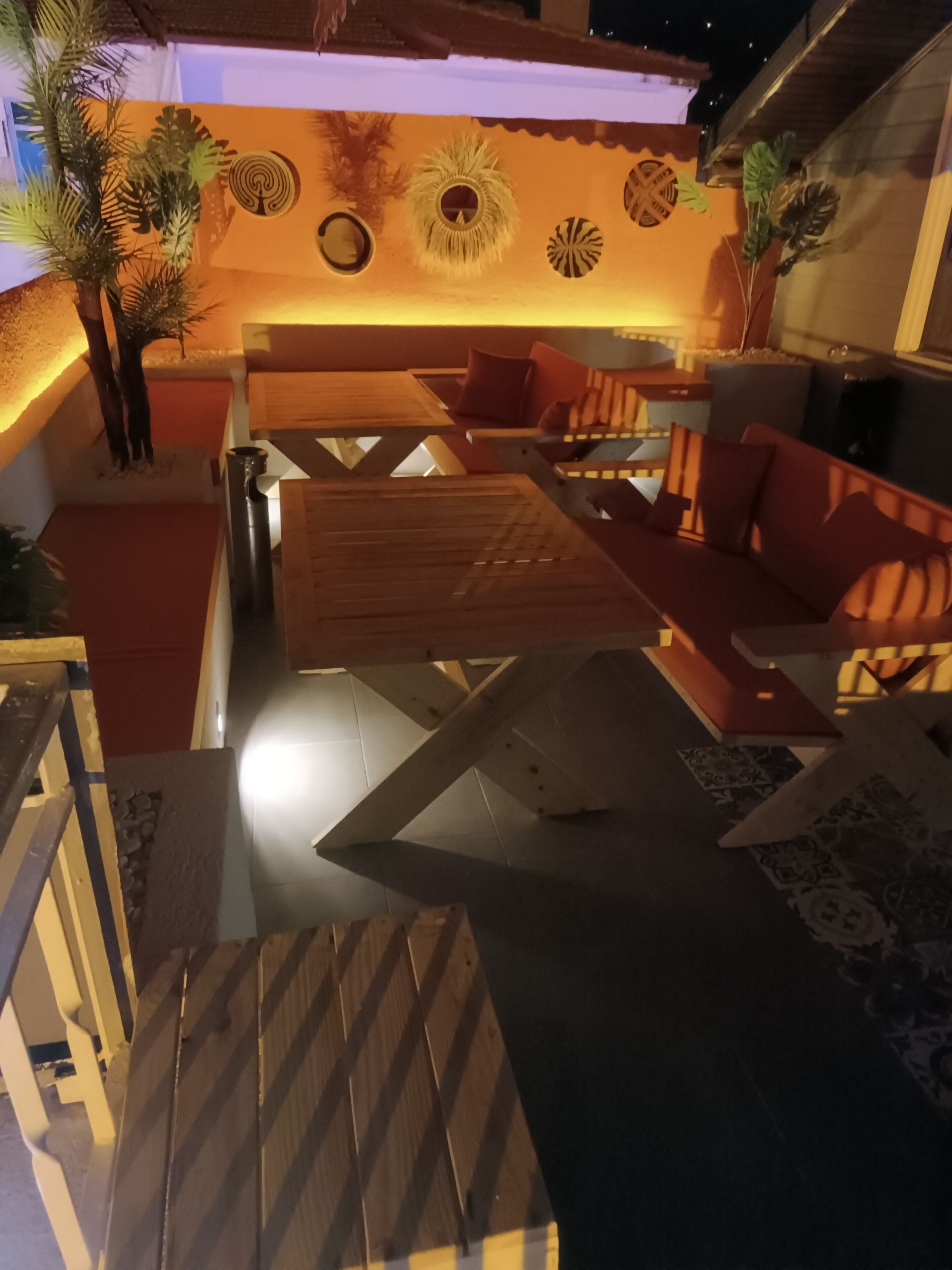 A modern outdoor seating area with a wooden table and red cushioned chairs, surrounded by decorative wall art and potted plants.