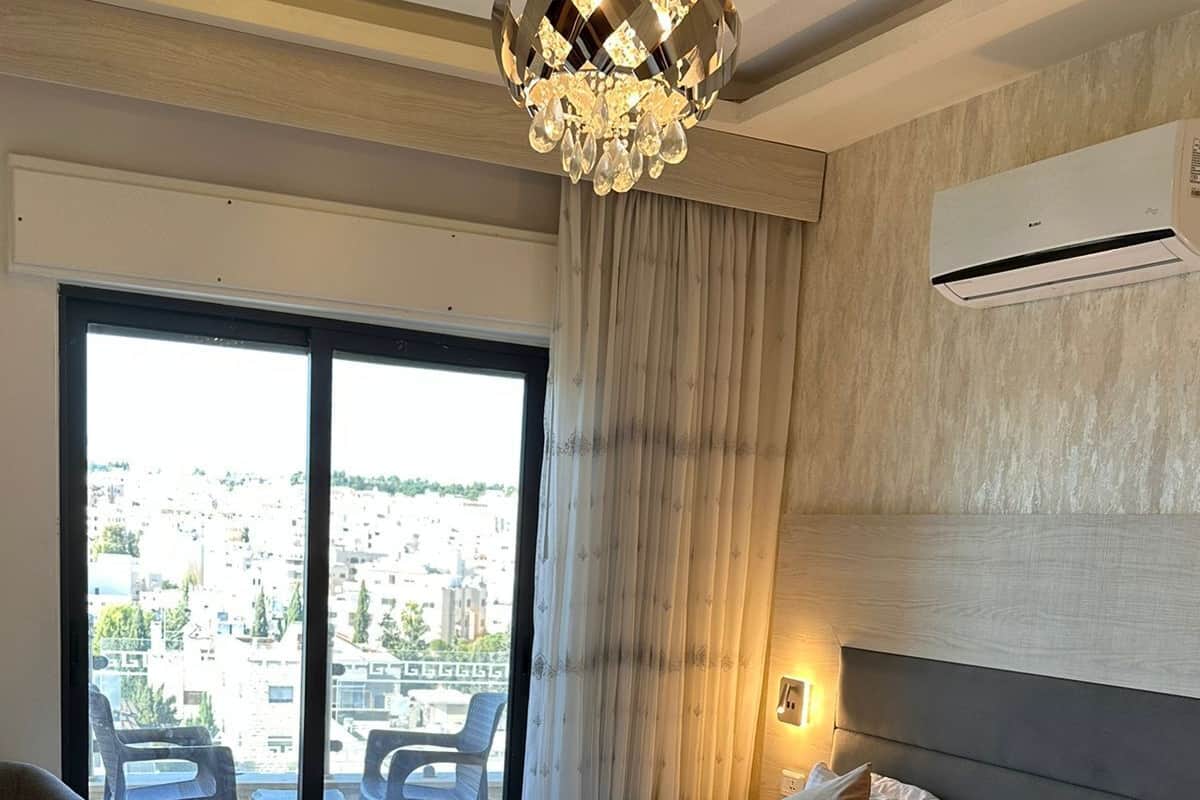 Hotellihuone, jossa on suuri sänky, ikkunasta avautuva näkymä kaupunkiin, kattokruunu ja ilmastointilaite.