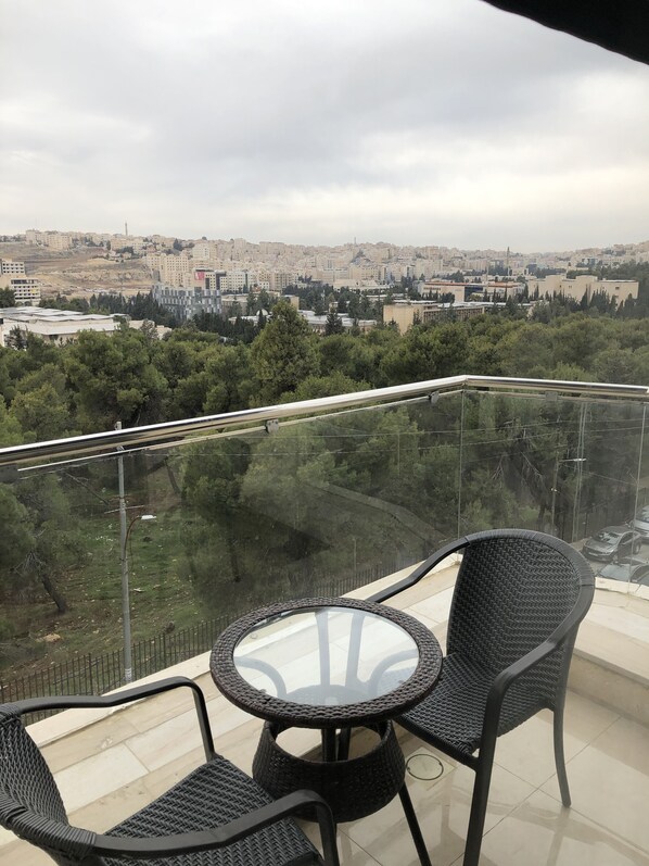 Uma varanda com mesa e cadeiras de vidro, com vista para um horizonte urbano com edifícios e árvores.