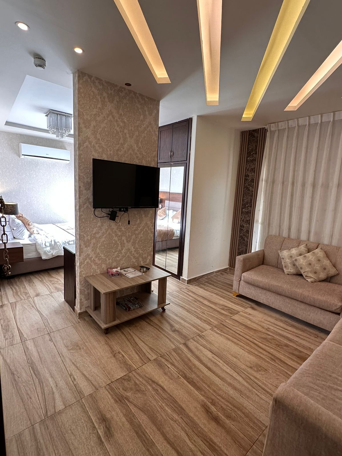 A modern living room with a flat-screen TV mounted on a wall, a wooden coffee table, a beige sofa, and a large window with curtains.