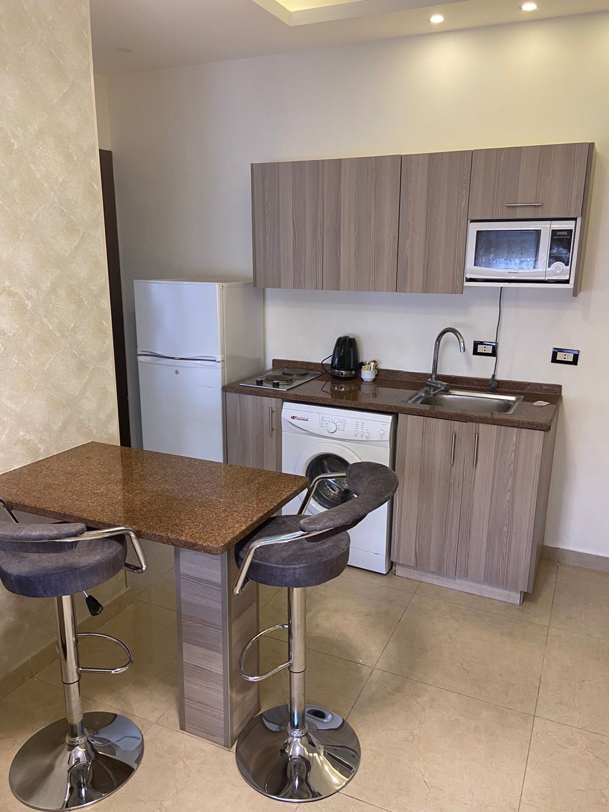 Una cocina compacta con microondas integrado, un lavabo y una lavadora. Hay un taburete de bar y una mesa con dos sillas.