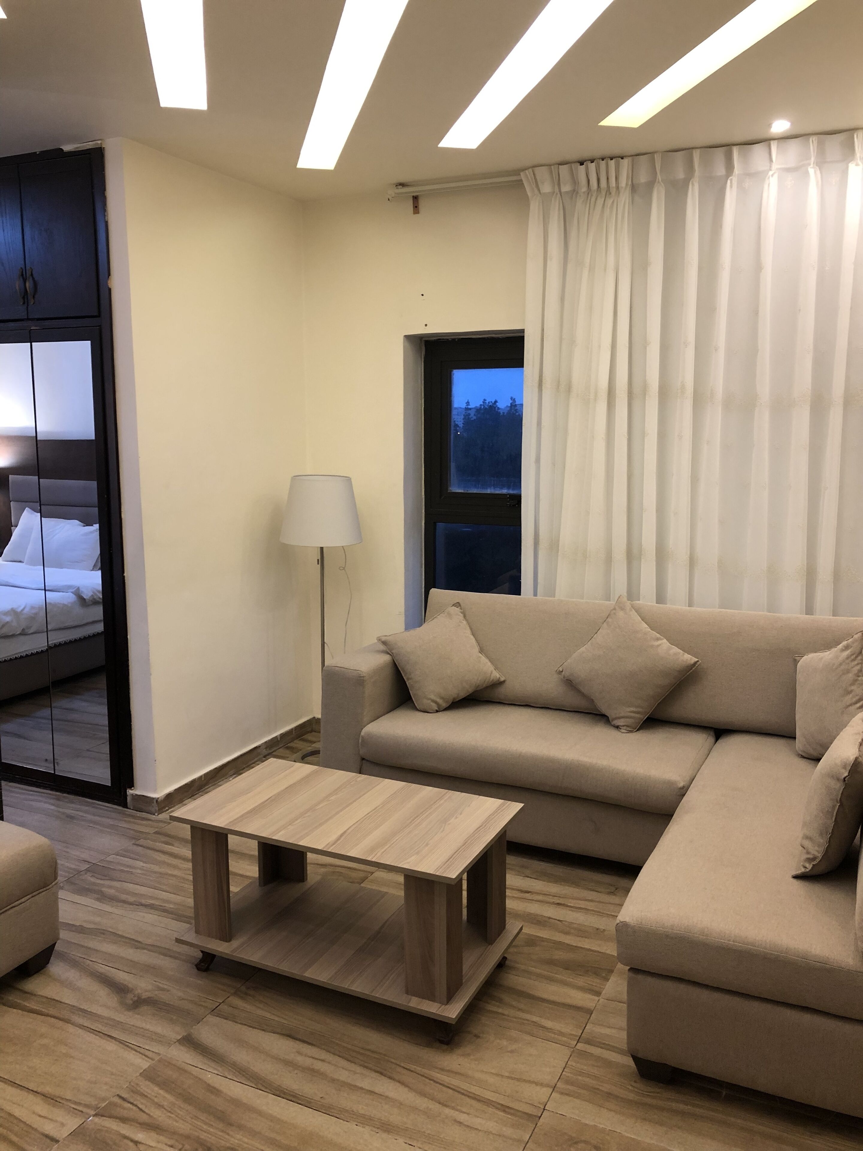 A modern living room with a sofa, a coffee table, and a lamp. There is a window with curtains and a view of trees outside.