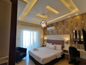 Quarto de hotel moderno com uma cama grande, uma mesa de trabalho e uma cadeira. Há um lustre, um ar condicionado e uma janela com cortinas.