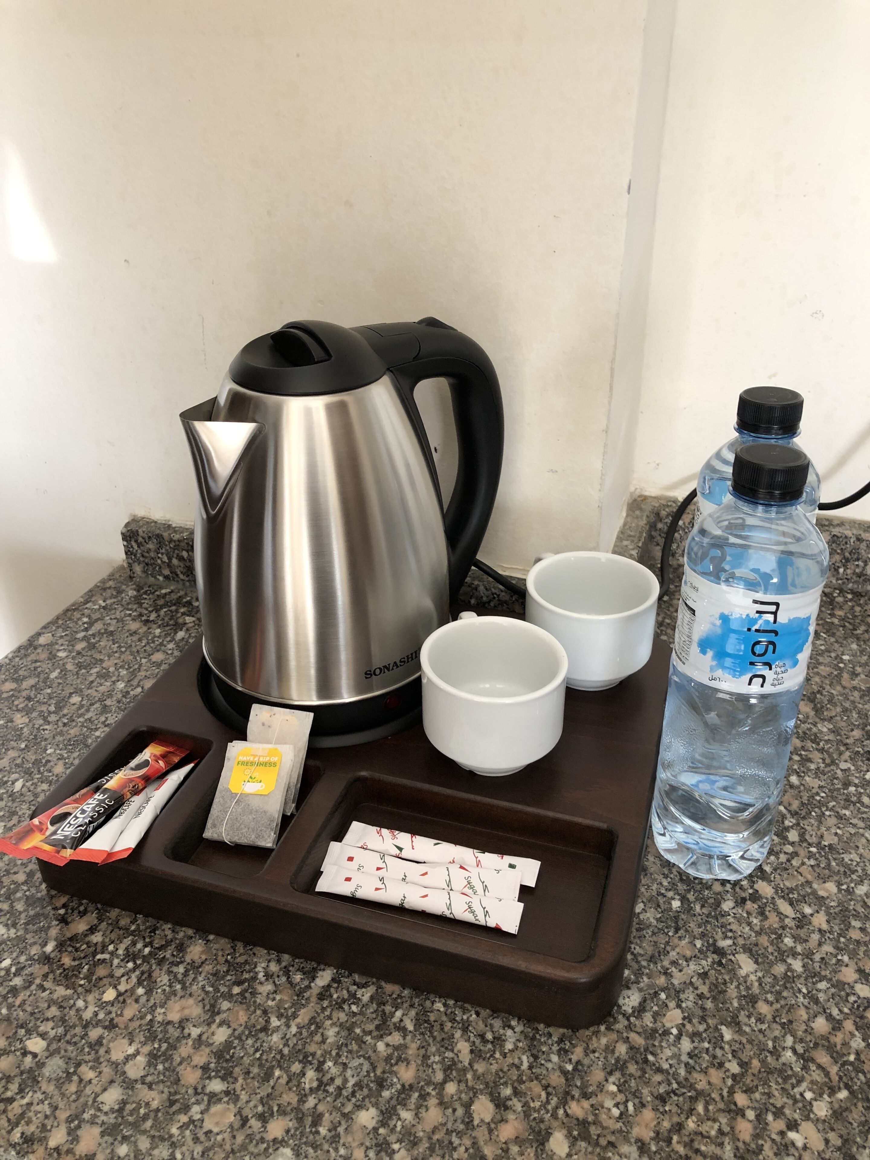 A kitchen area with a stainless steel kettle, two white cups, a water bottle, and a tray with tea bags and a packet of instant coffee.