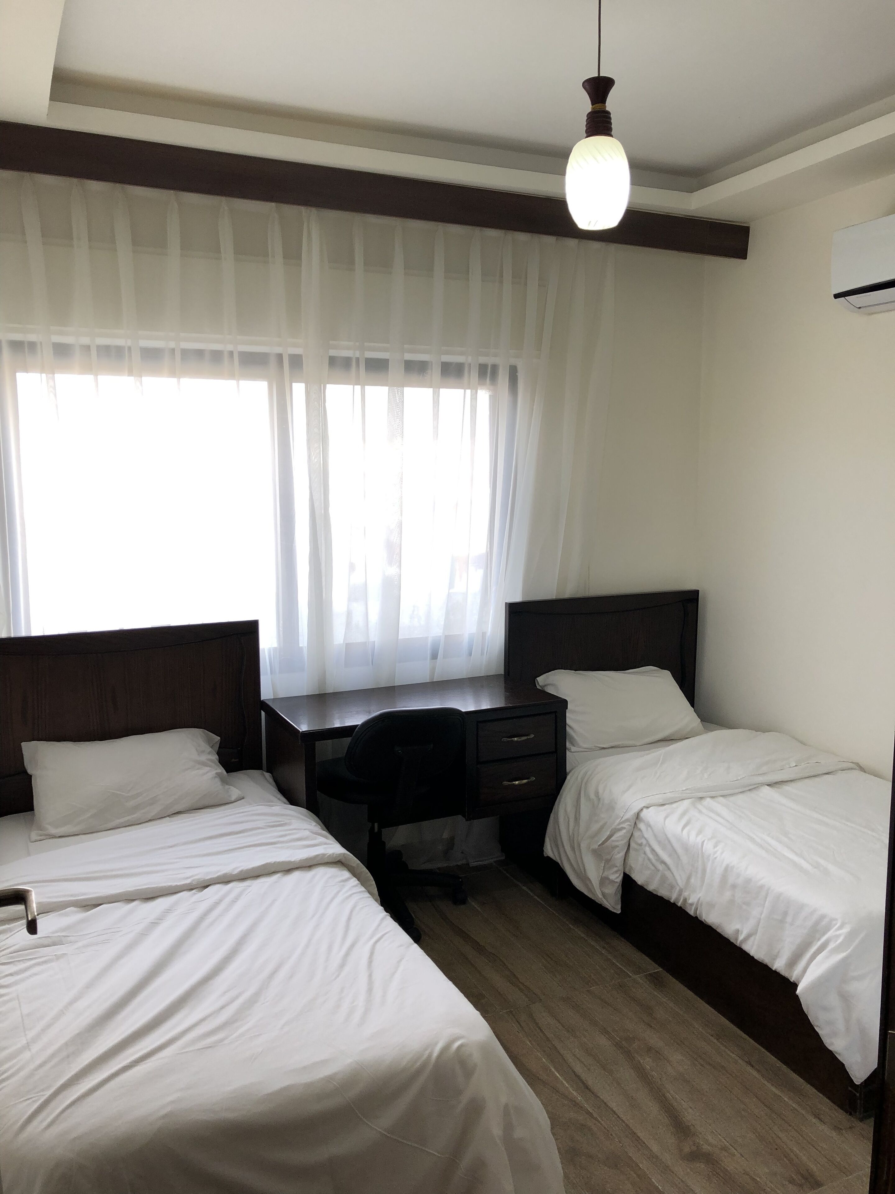 A hotel room with two single beds, a desk with a chair, and a large window with sheer curtains.