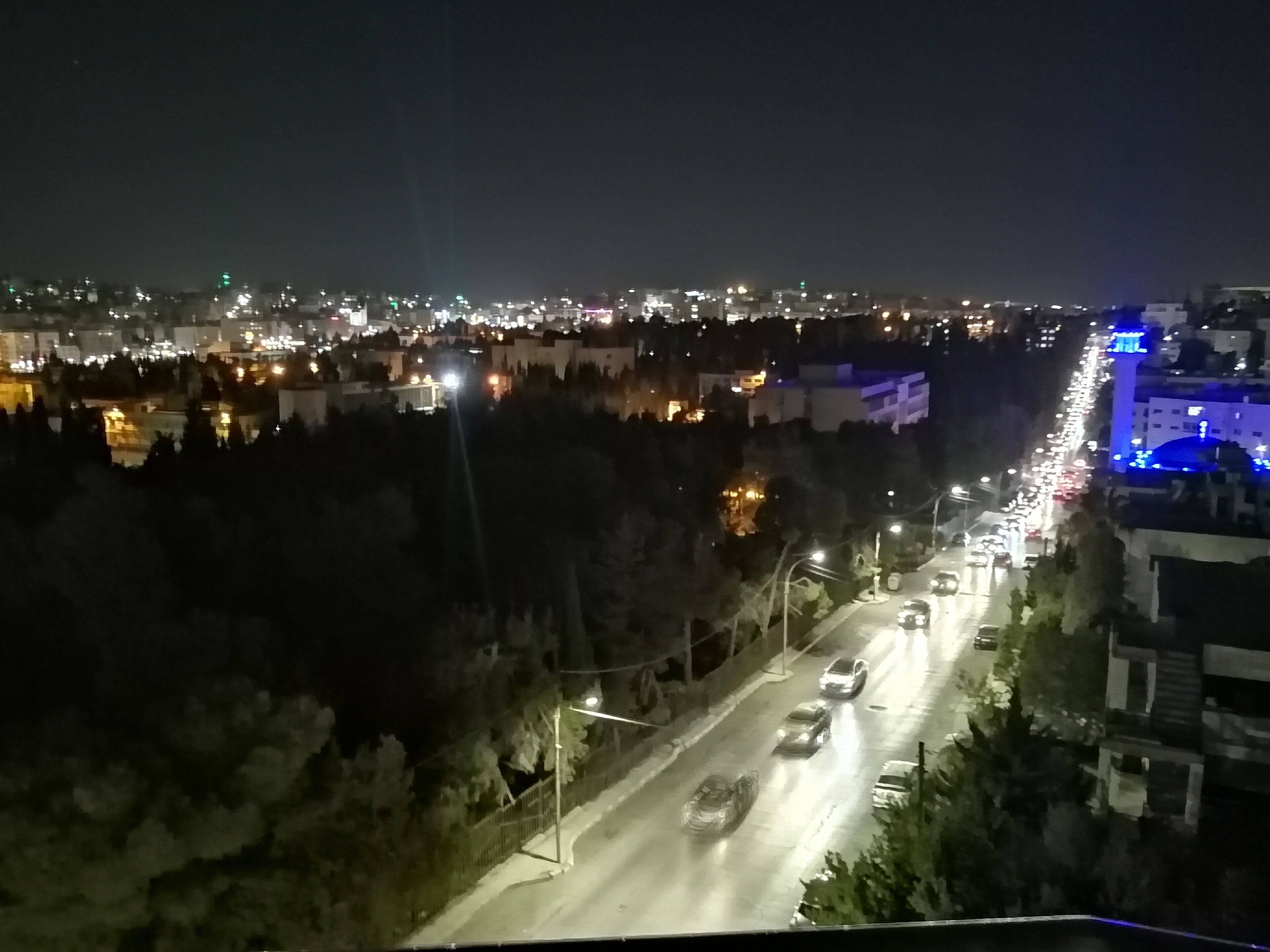 A nighttime cityscape with a busy road, illuminated buildings, and a distant view of a city skyline.
