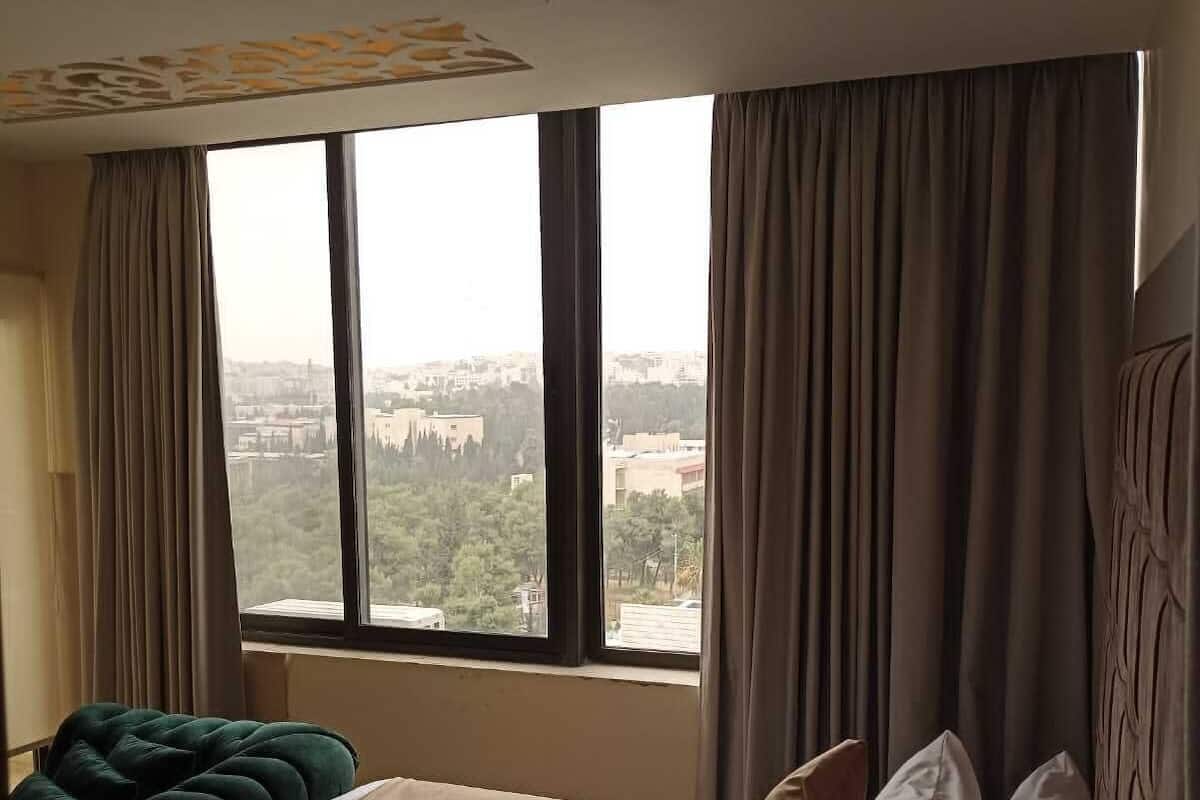 Hotellihuone, jossa on suuri ikkuna, valkoisilla lakanoiden peitetty sänky, vihreäverhoiltu penkki ja näkymä kaupunkimaisemaan.