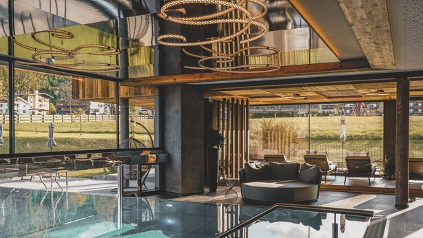 Un área de piscina interior moderna con paredes de vidrio, una sala de estar con asientos y vistas al exterior.