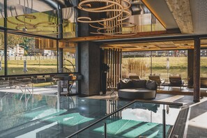 Un área de piscina interior moderna con paredes de vidrio, una sala de estar con asientos y vistas al exterior.