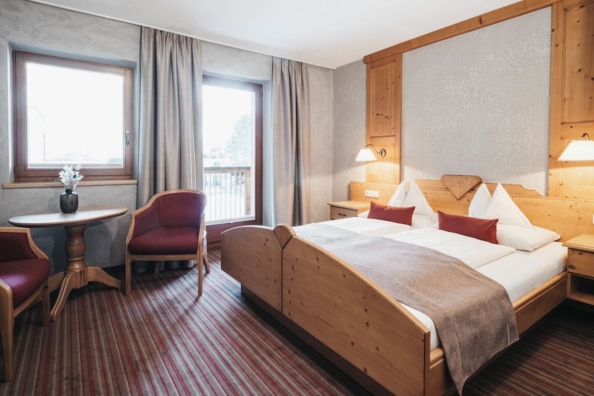 Une chambre d’hôtel avec un grand lit, une tête de lit en bois, une petite table avec un vase, une chaise et une fenêtre avec des rideaux.