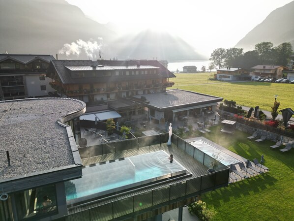 Un hotel moderno con piscina en la azotea, un espacio al aire libre cubierto y vistas a un lago y montañas al fondo.