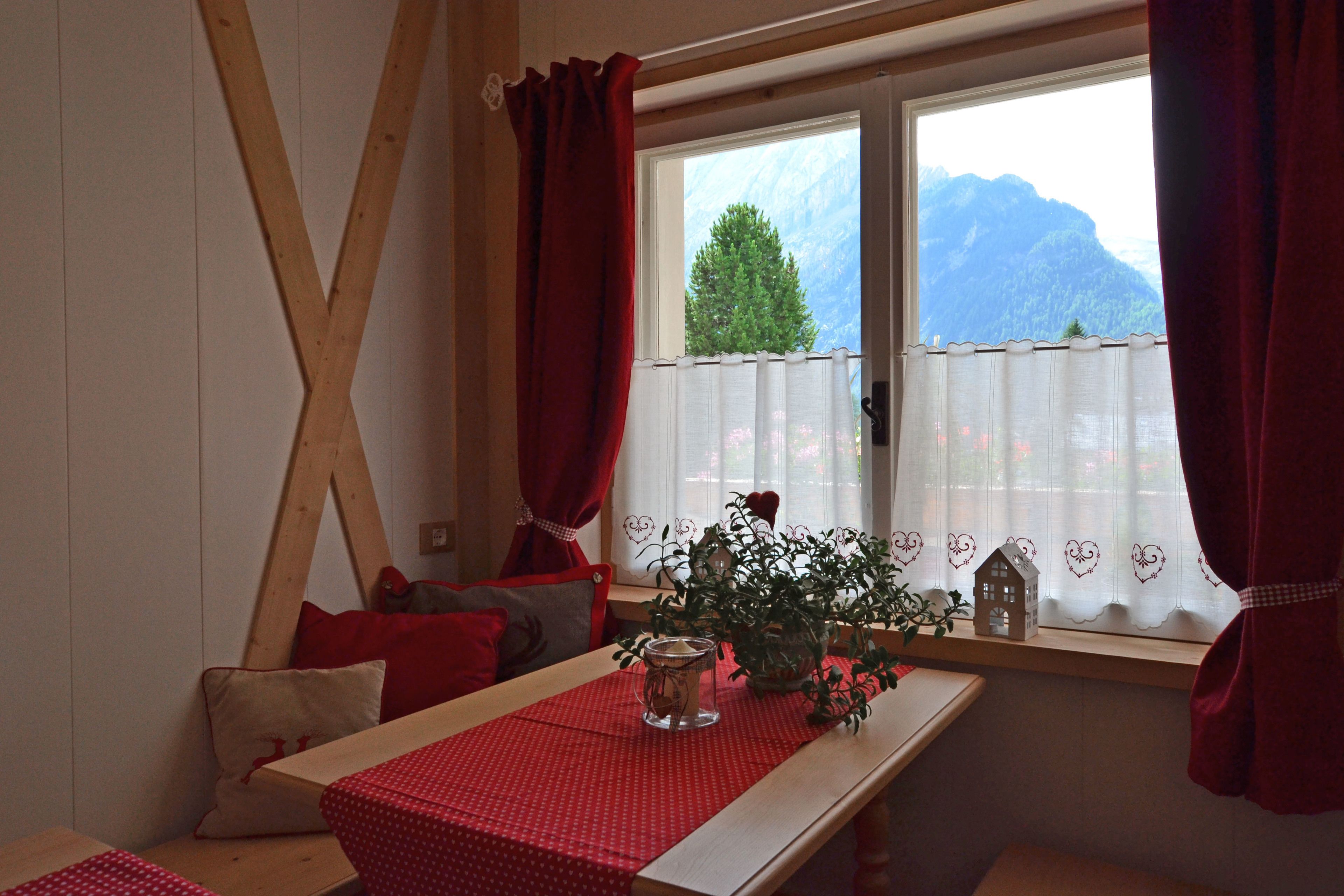 A cozy room with a wooden bench, a table with a red tablecloth, a potted plant, and a view of mountains through a window with red curtains.