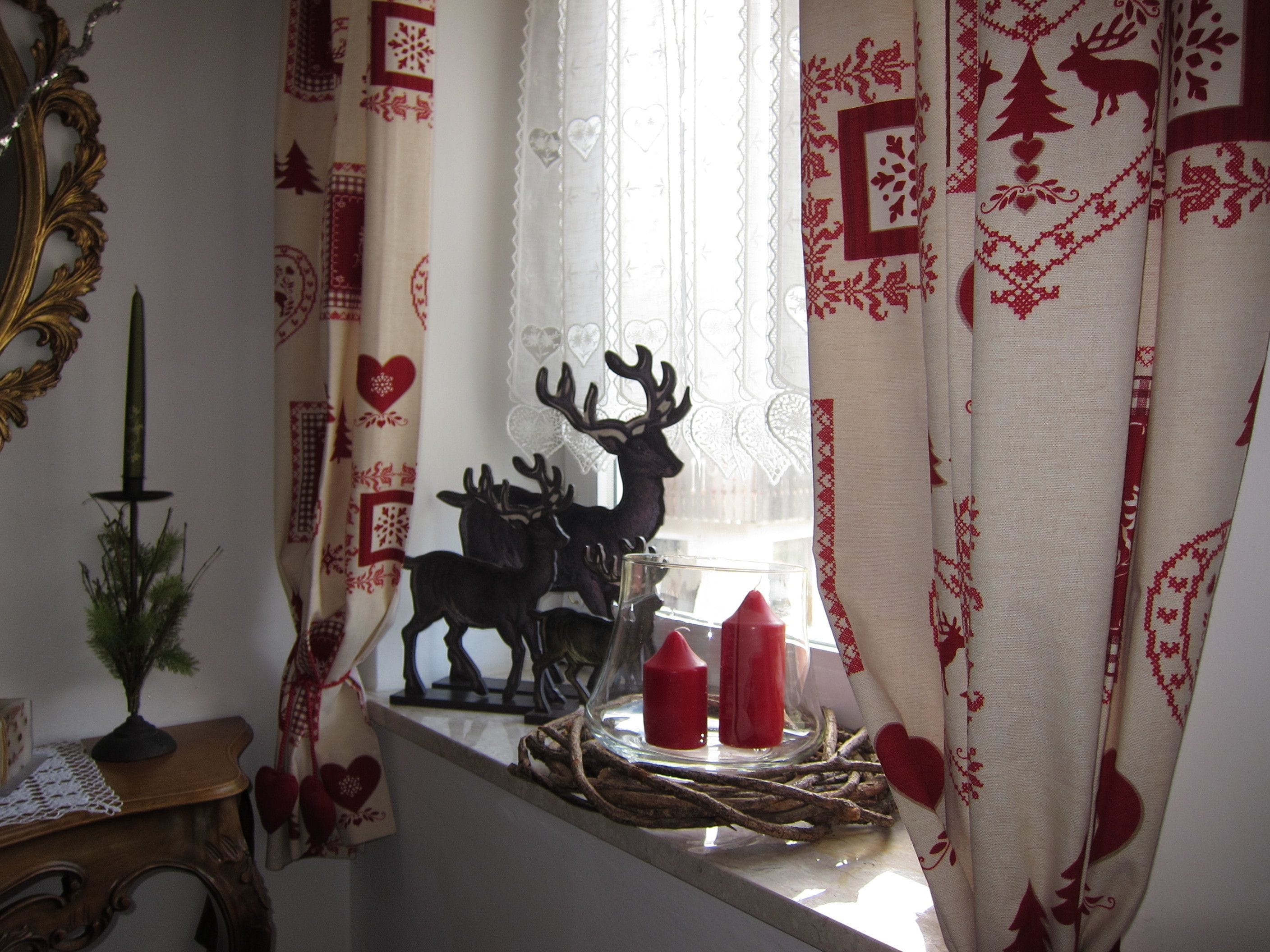 Una habitación con una ventana decorada con cortinas estampadas, una figurita decorativa de ciervo y dos velas rojas sobre un repisa de madera.