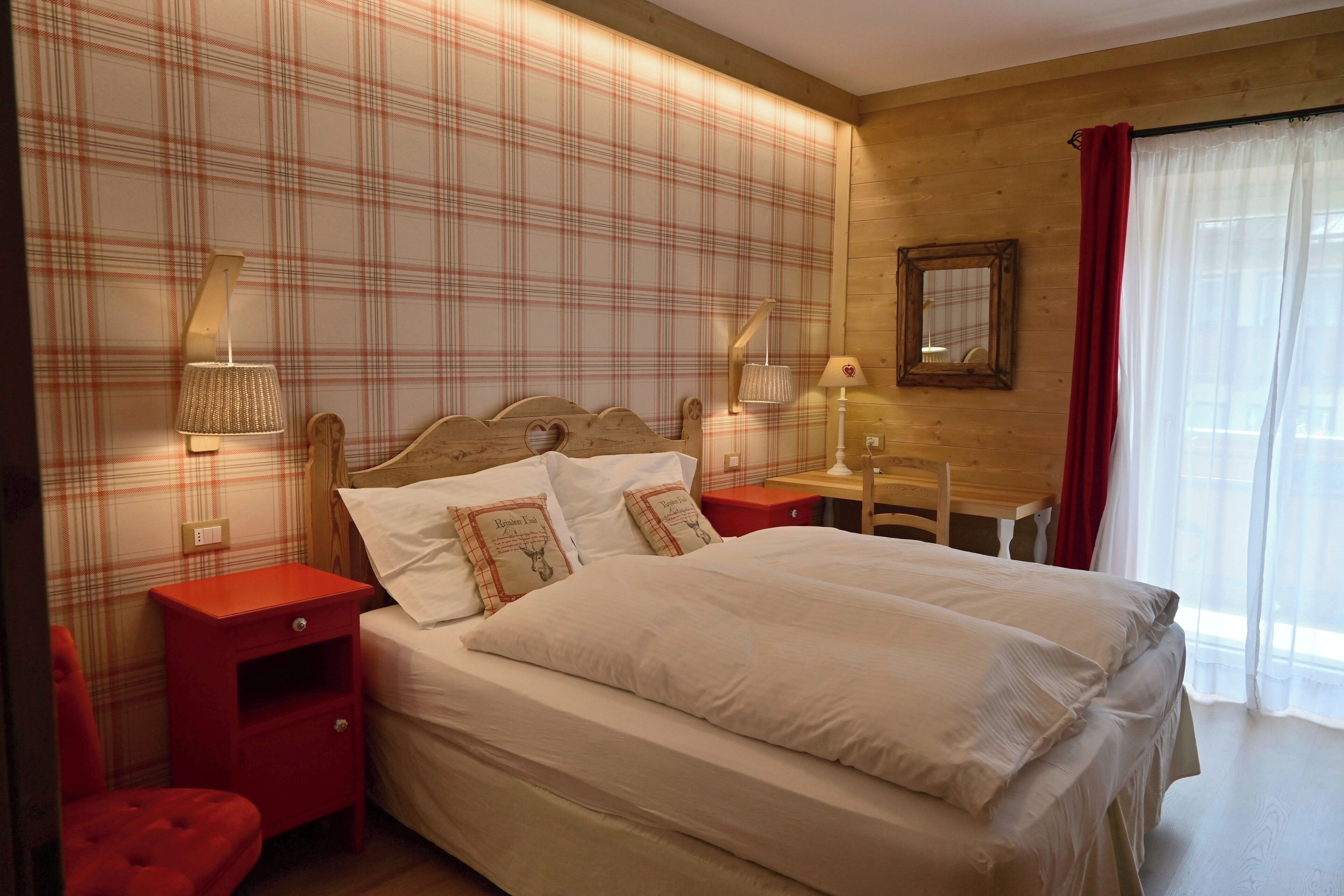 Cama bem arrumada com lençóis brancos e almofadas decorativas, cabeceira de madeira, duas mesas de cabeceira com abajures, uma escrivaninha de madeira pequena e uma cadeira vermelha.