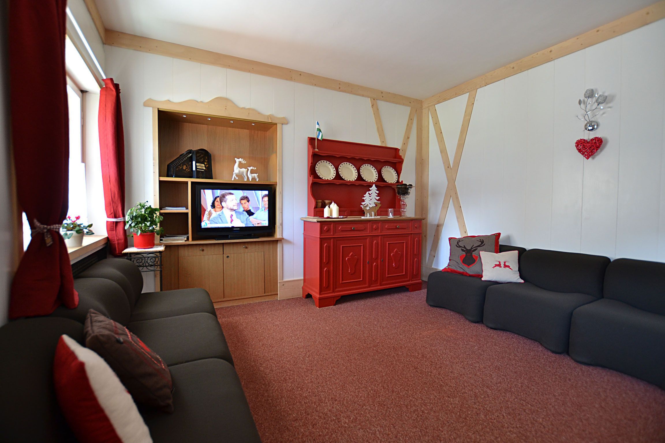 A living room with a dark sofa, a red cabinet with decorative items, a television, and a wooden ceiling structure.