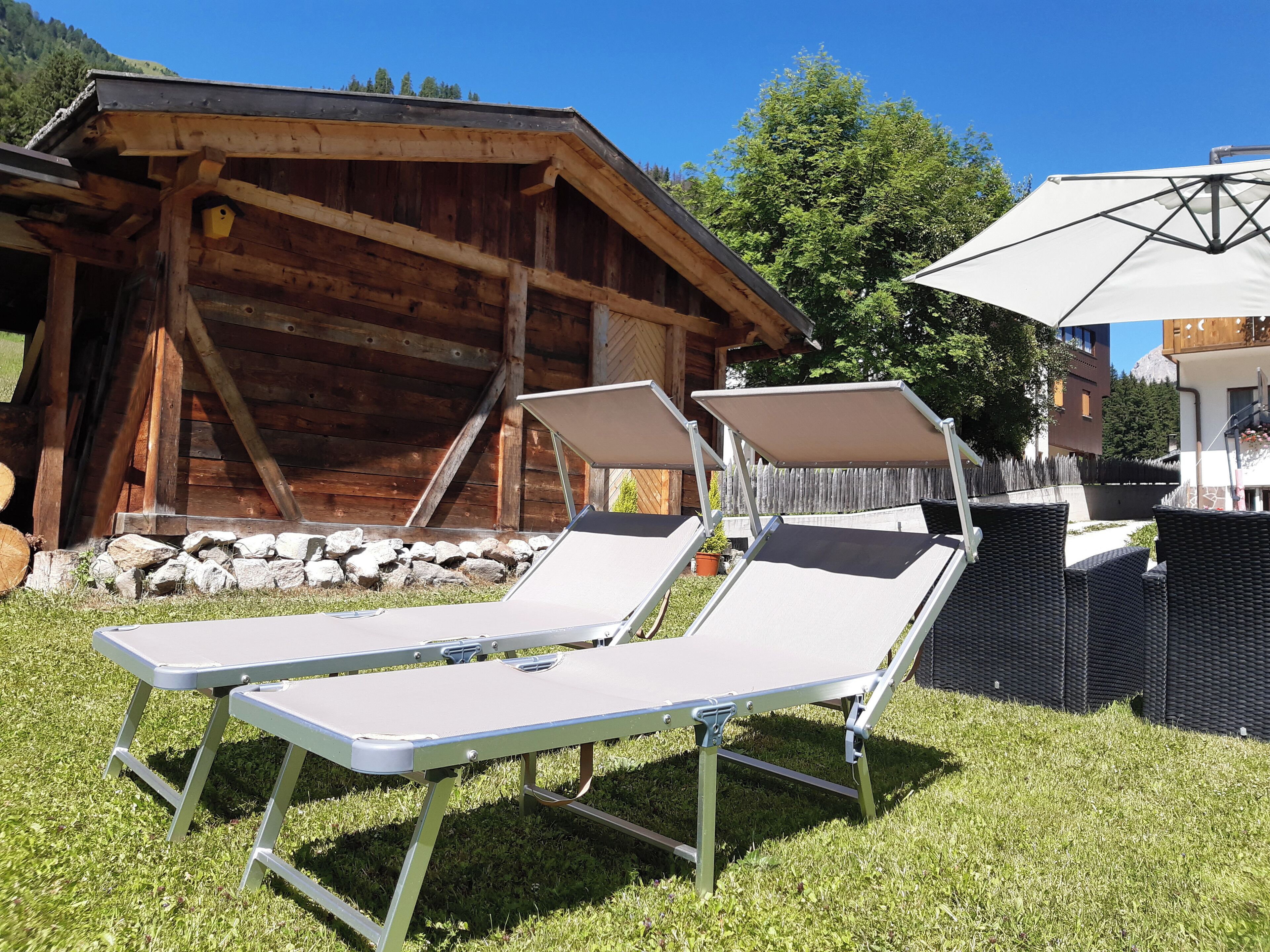 Two lounge chairs with a table, a wooden cabin, and a white umbrella in a grassy area.