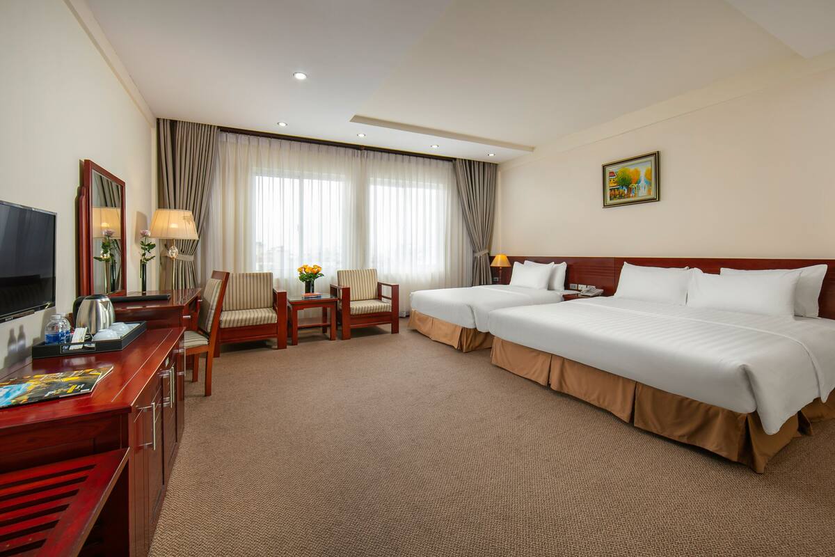 A hotel room with a large bed, a desk with a TV, a chair, and a small table with a lamp and flowers.