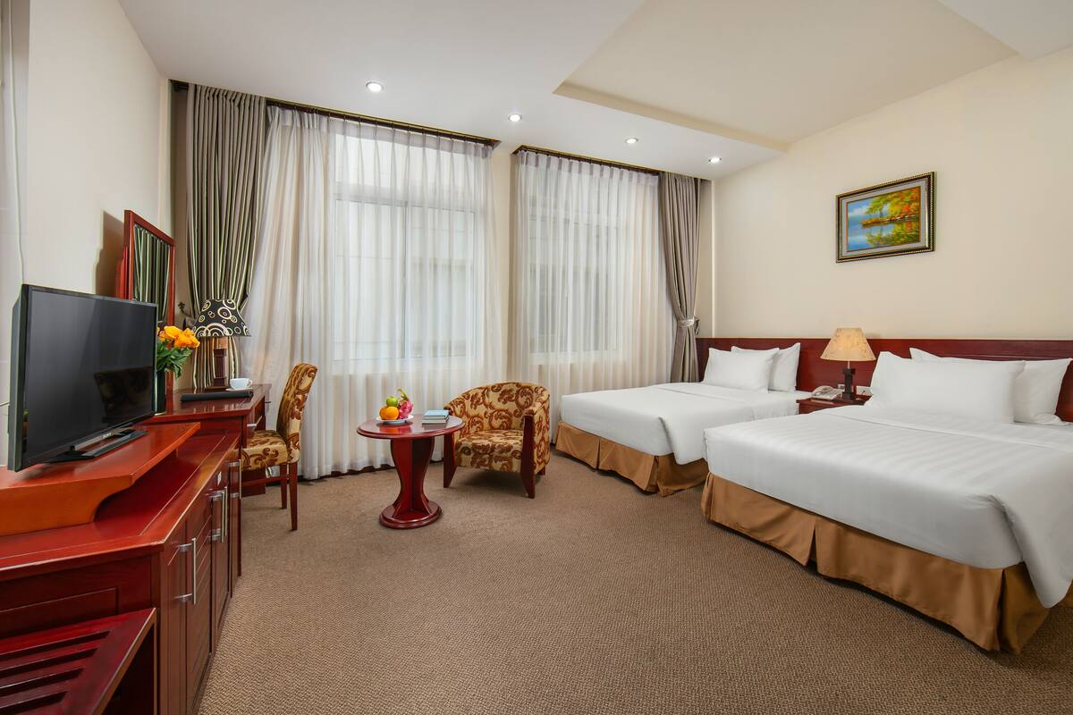 A hotel room with two beds, a TV, a desk with a chair, a small table with fruit, and curtains.
