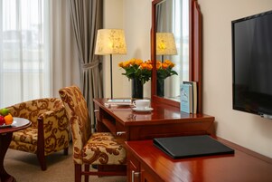 A hotel room with a wooden desk, a chair, a vase of flowers, a lamp, a television, and a fruit bowl.
