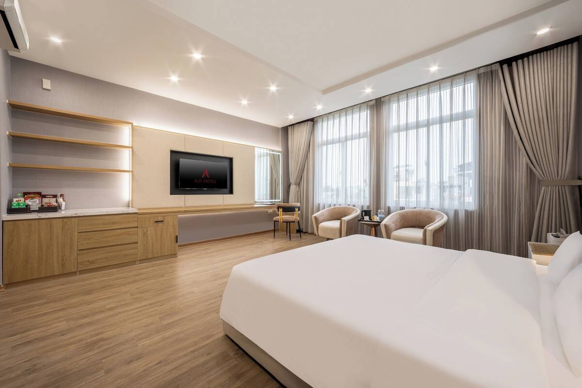 A modern hotel room with a large bed, a flat-screen TV mounted on the wall, a desk with a chair, and a seating area with two chairs.