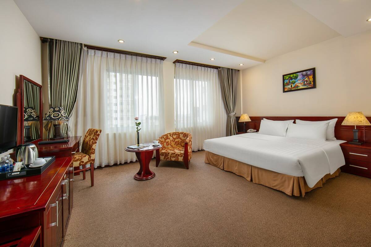 A hotel room with a bed, a desk with a television, a chair, a small table with a vase, and curtains.