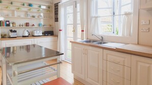 Une cuisine avec des meubles blancs, un plan de travail en acier inoxydable, un évier à deux bacs et une fenêtre avec des rideaux.