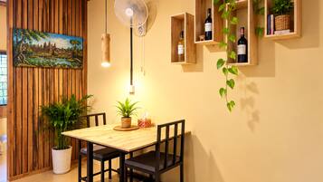 Un comedor con una mesa y sillas de madera, un ventilador de pared, y estantes con botellas y plantas.