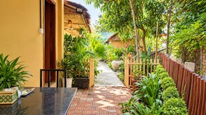 A courtyard with a brick pathway leading to a wooden door, surrounded by lush greenery and a thatched-roof structure. - Siem Reap Homesteading (Siem Reap)