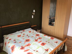 Um quarto com uma cama, um guarda-roupa de madeira, uma mesa de cabeceira e um abajur fixado na parede.