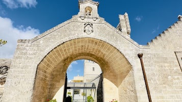 An arched stone gateway leading to a building with a bell tower, potted plants, and a clear blue sky.