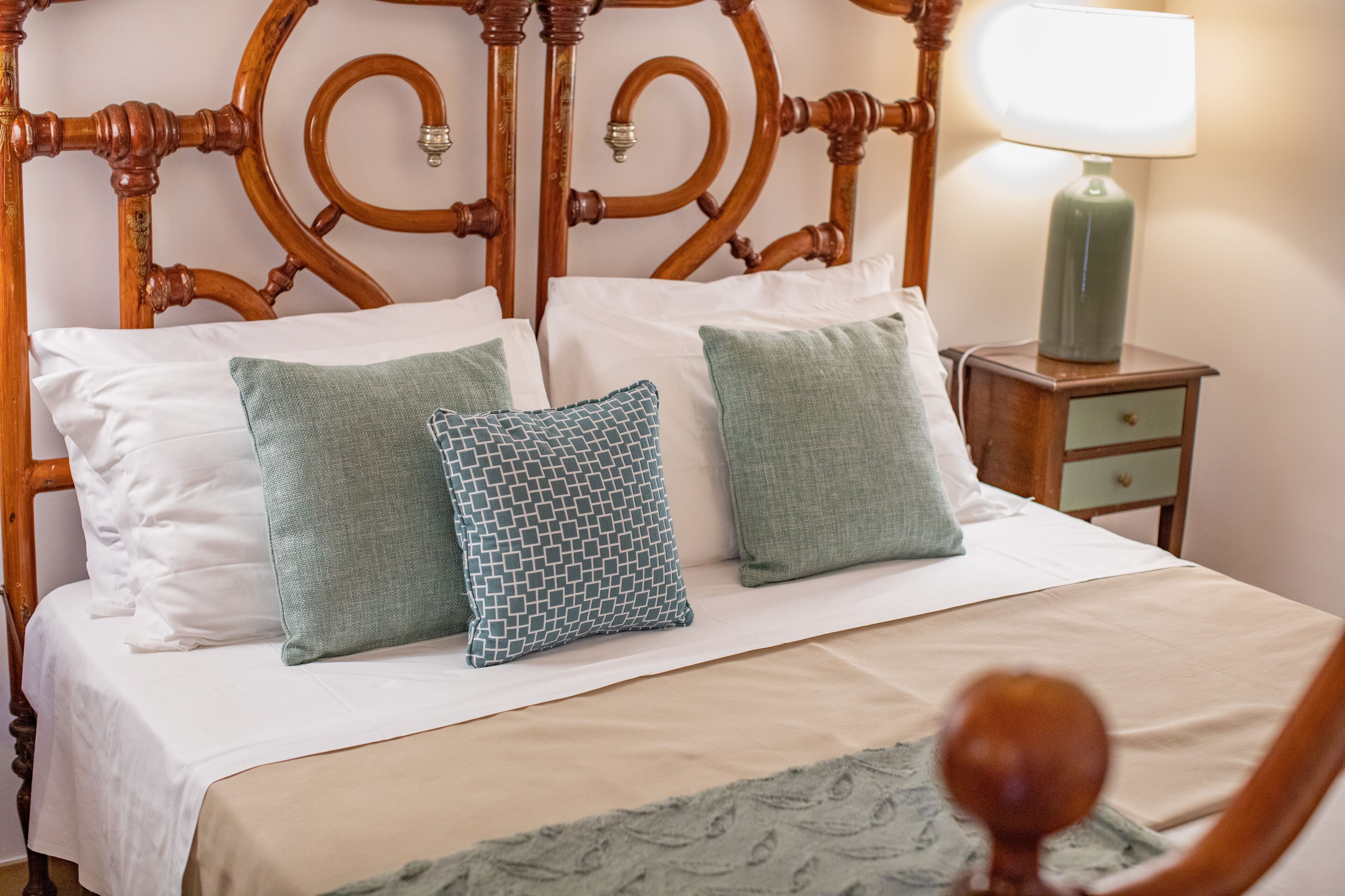 Una cama bien hecha con tablero de madera, dos almohadas, una mesita de noche con lámpara y un cajón pequeño.