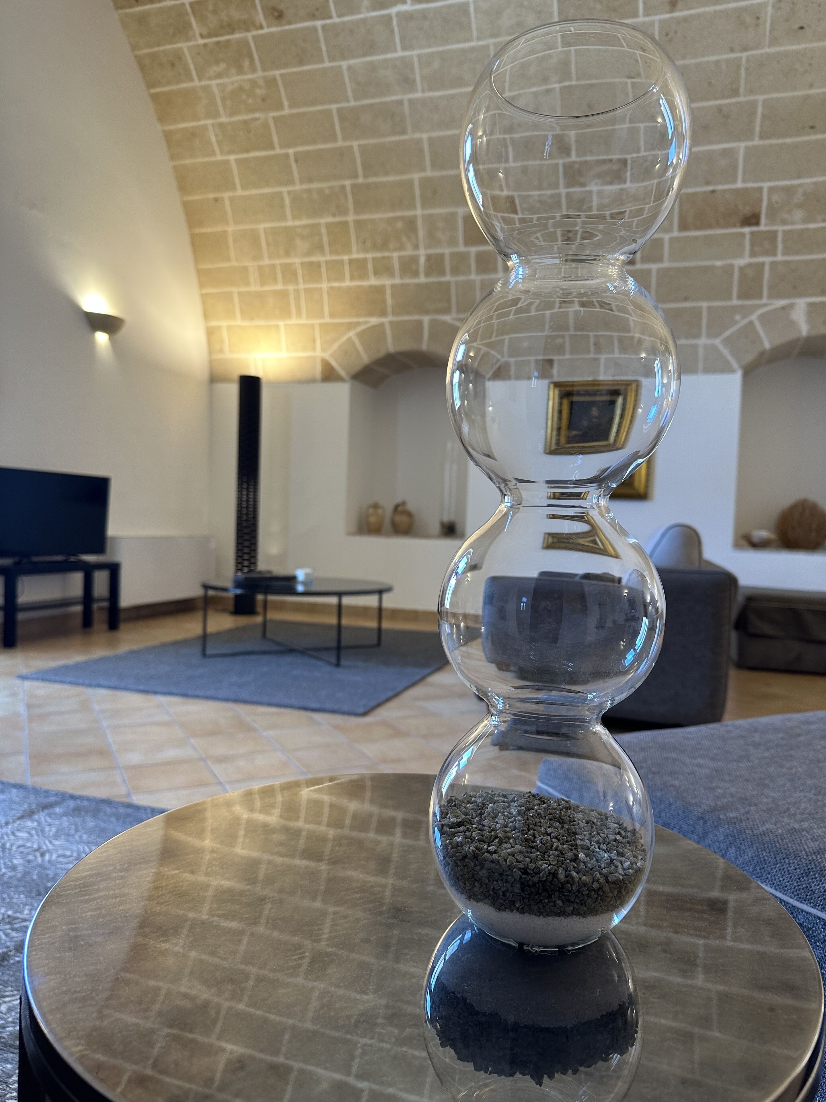 Una ampulleta de vidrio transparente con arena sobre una superficie reflectante en una habitación con piso de baldosas, chimenea y un cuadro en la pared.