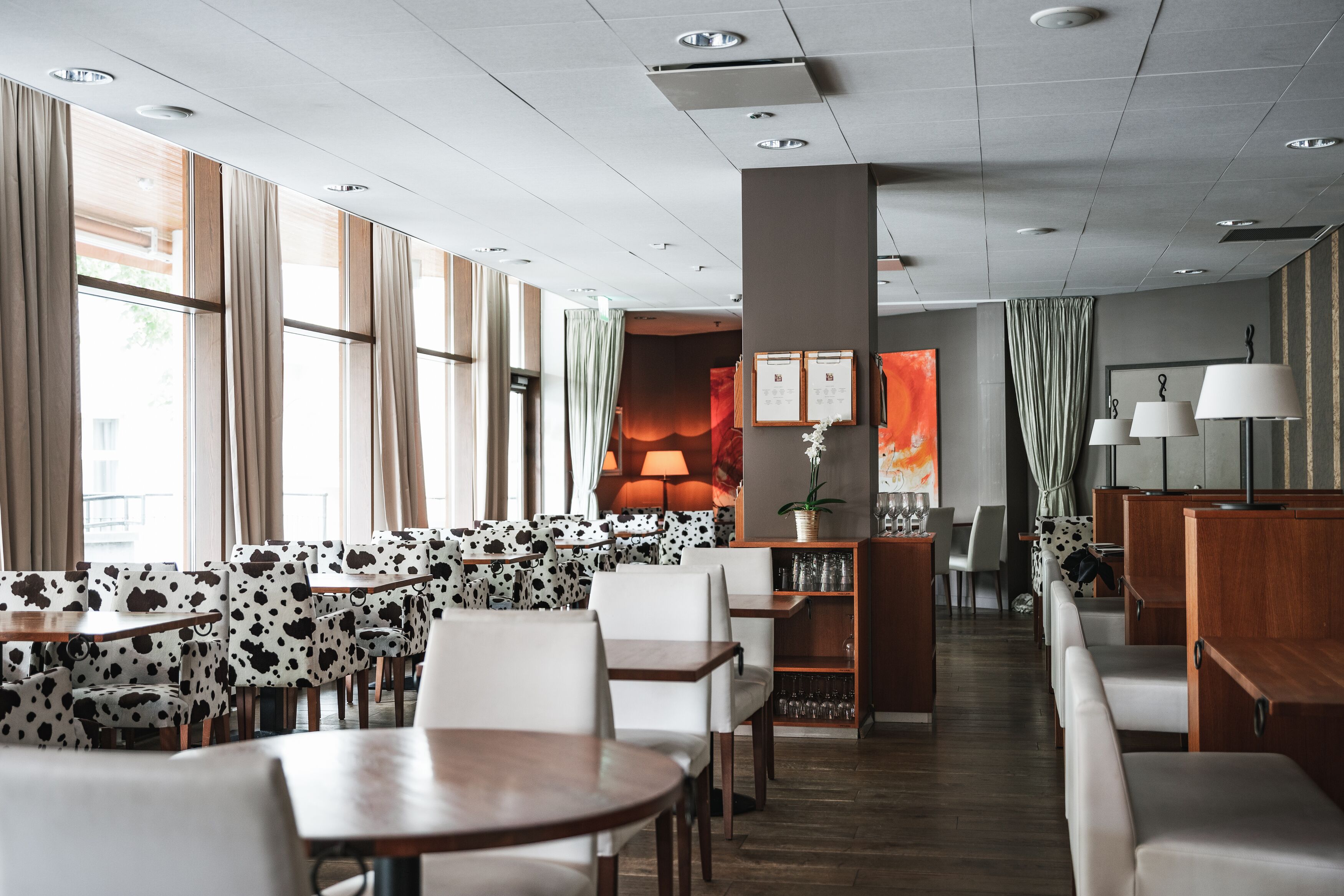 A modern hotel lobby with a seating area featuring cow-print upholstered chairs and tables, a central fireplace, and a view of the outdoors through large windows.