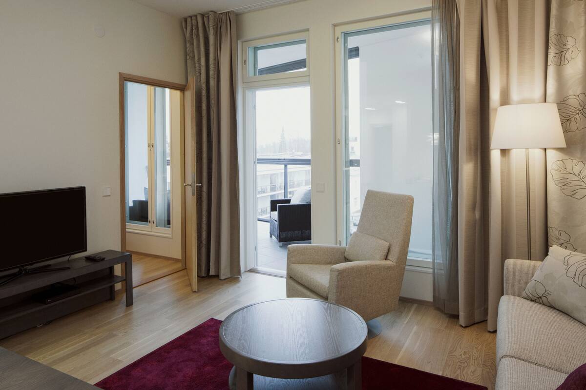 Une chambre d’hôtel moderne équipée d’un téléviseur à écran plat, d’une table basse ronde, d’un canapé et d’un fauteuil. Une grande fenêtre, agrémentée de rideaux, offre une vue sur le paysage urbain.
