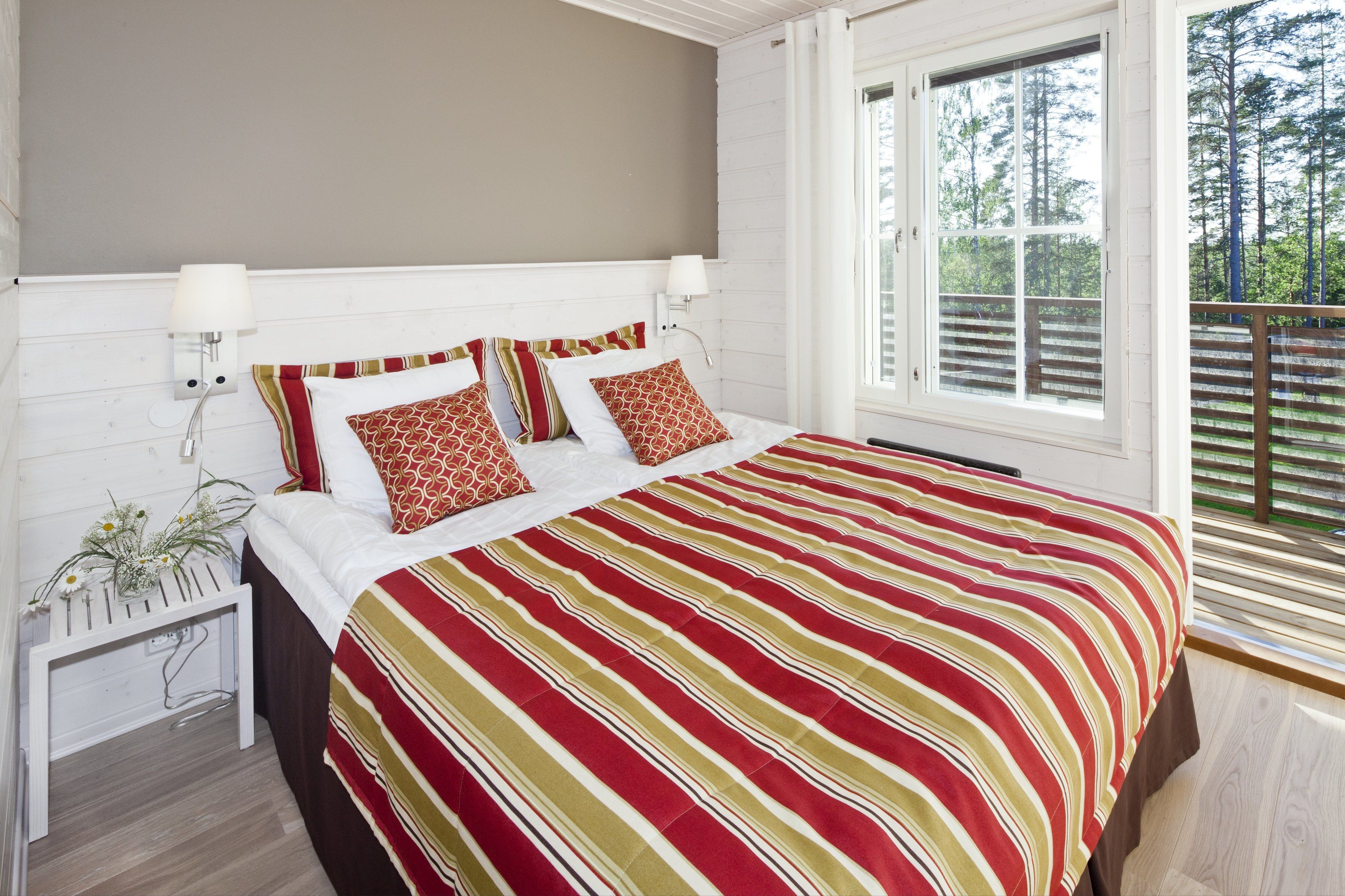 A bedroom with a bed, two bedside lamps, a nightstand with a plant, and a window with a view of trees.