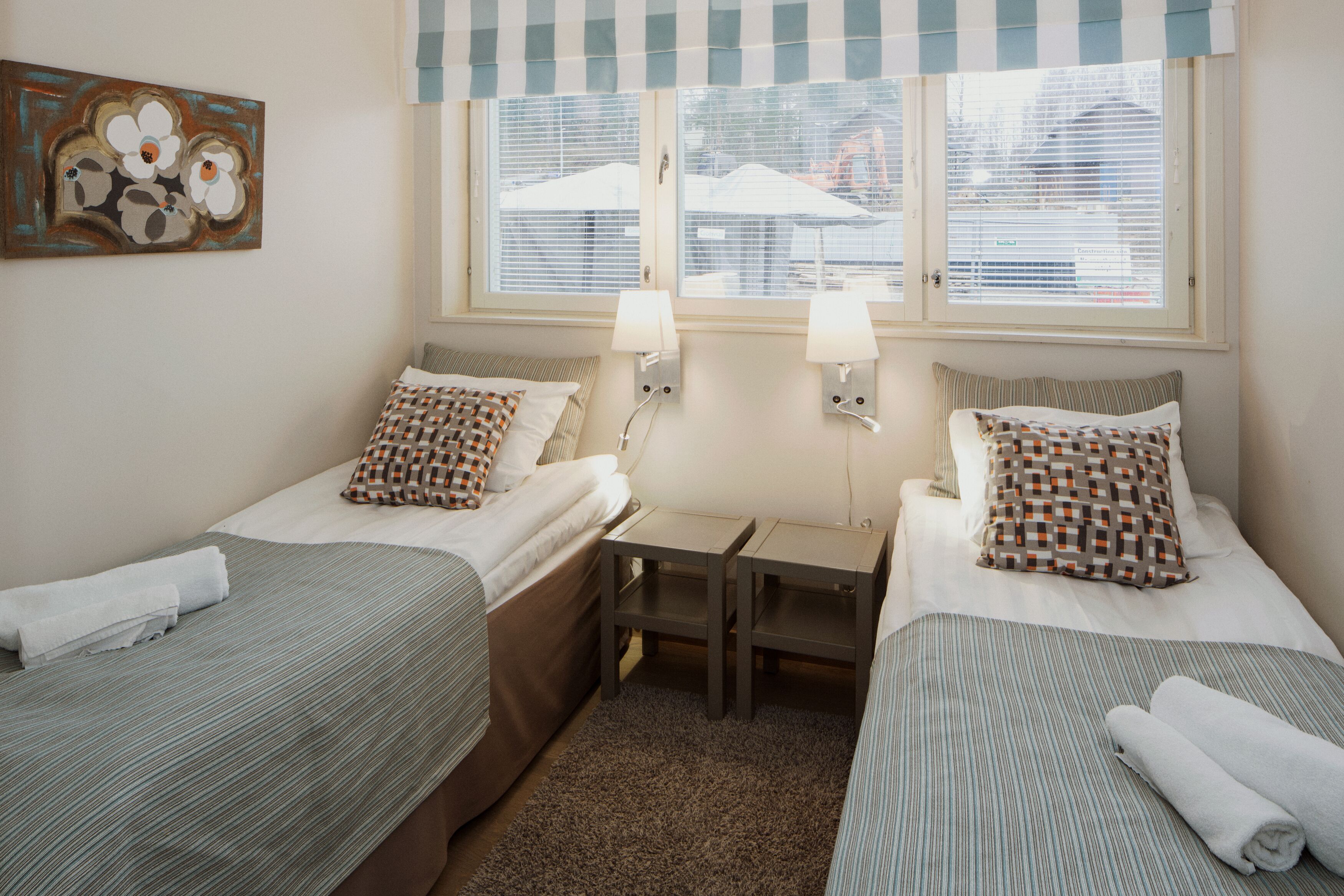 A twin room with two beds, a small table, two wall-mounted lamps, and a window with blinds.
