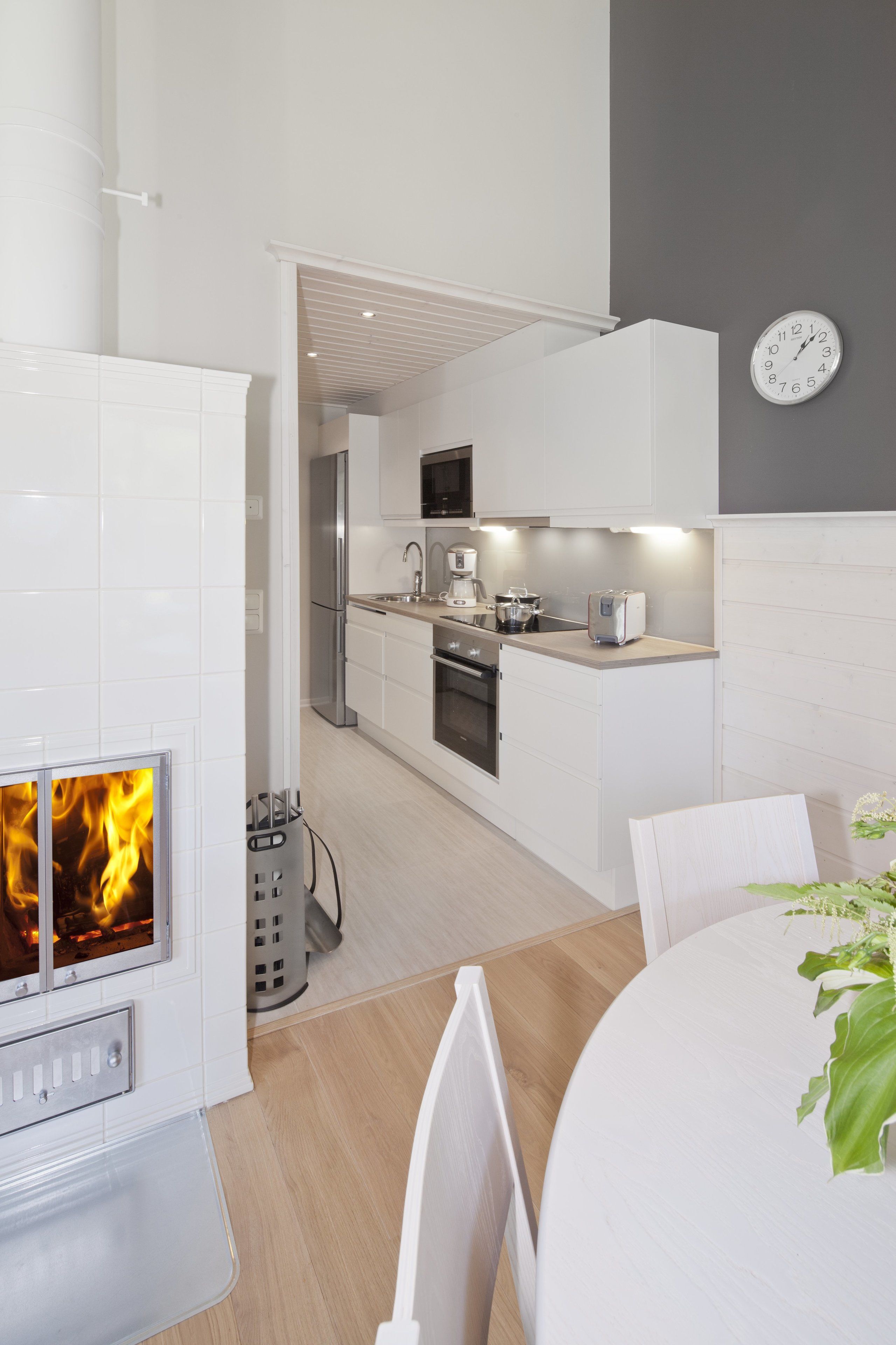 A modern kitchen with white cabinetry, a built-in oven, and a microwave. There is a dining area with a table and chairs, and a clock on the wall.