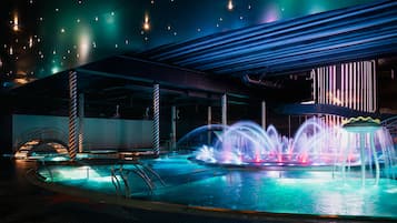 A nighttime scene of a modern building with a pool area featuring illuminated water jets and a unique architectural design with a sloped roof and vertical light strips.