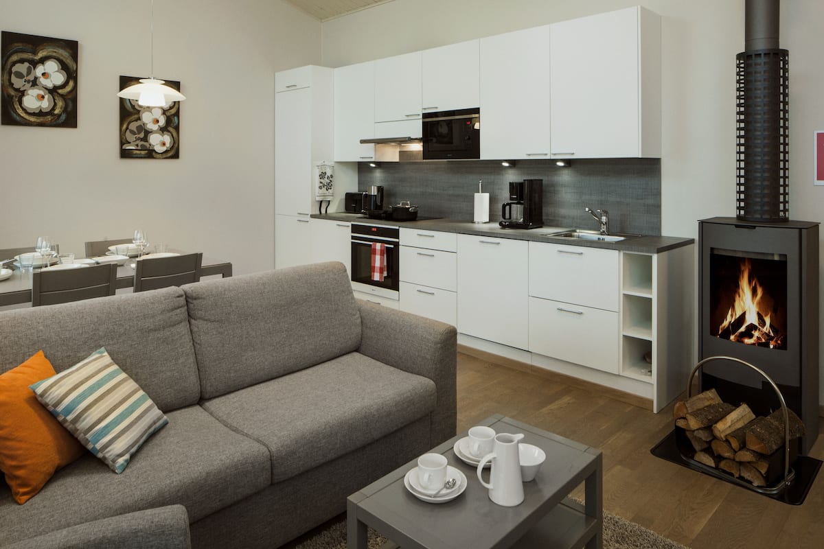Un salon moderne avec un canapé gris, une table basse avec des tasses et une bouilloire, et une cuisine dotée de meubles blancs et d’un poêle à bois.