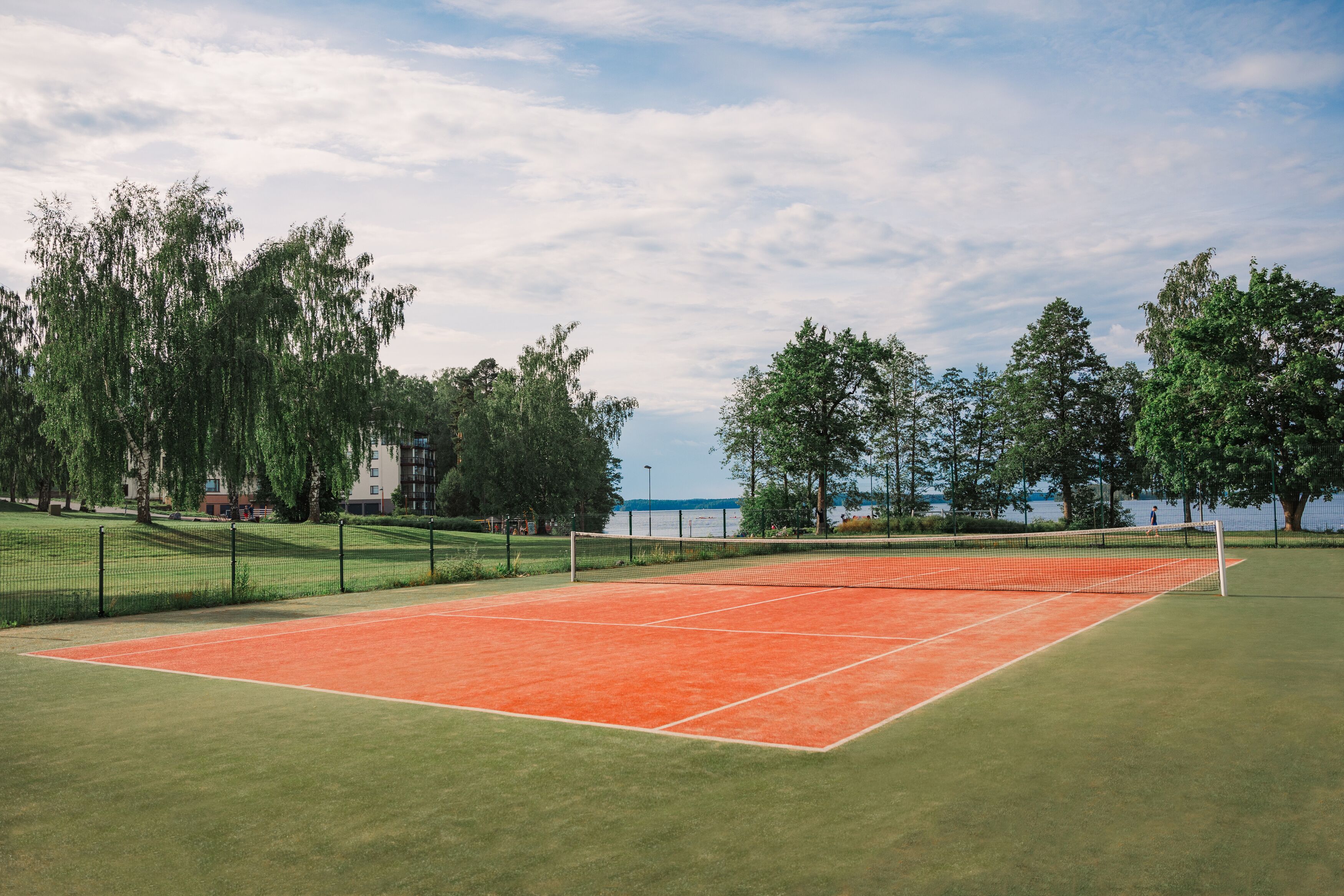 An outdoor tennis court with a net, surrounded by trees and a body of water in the background.