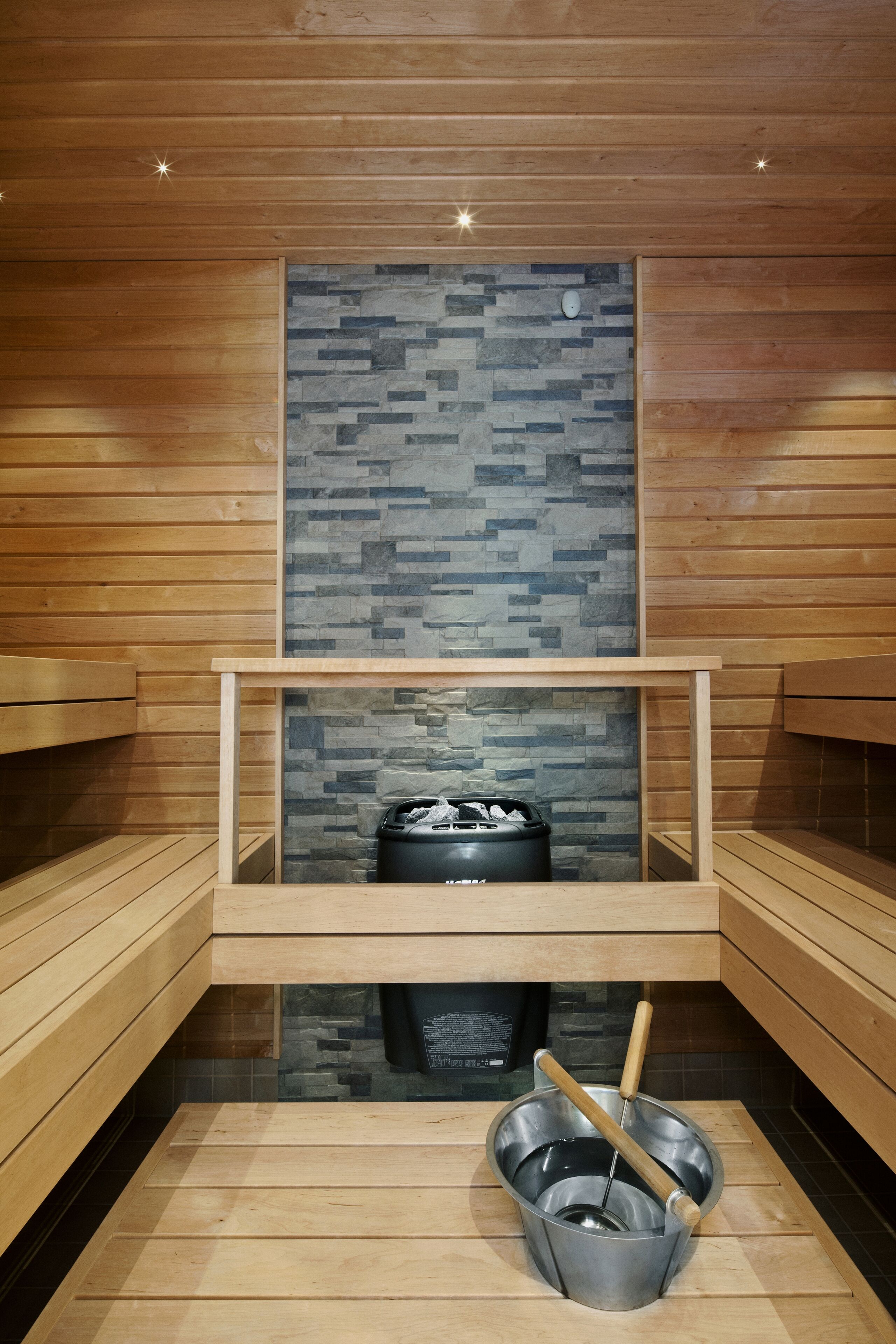 A sauna with wooden benches, a stone-tiled wall, and a metal bucket with a ladle.
