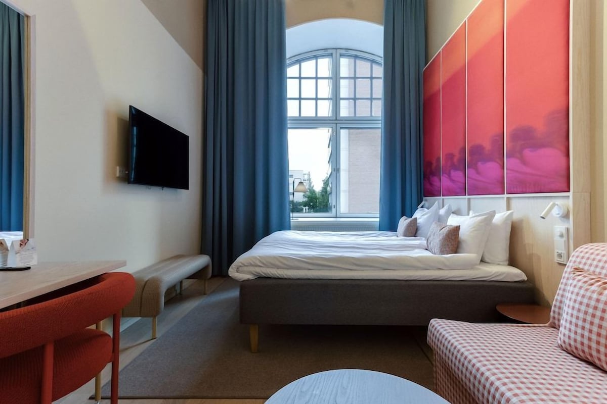 Habitación de hotel con cama, escritorio, silla, televisor y ventana con cortinas.