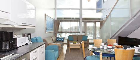 A modern kitchen with white cabinets, a dining area with a glass table, blue chairs, and a sofa with teal cushions.